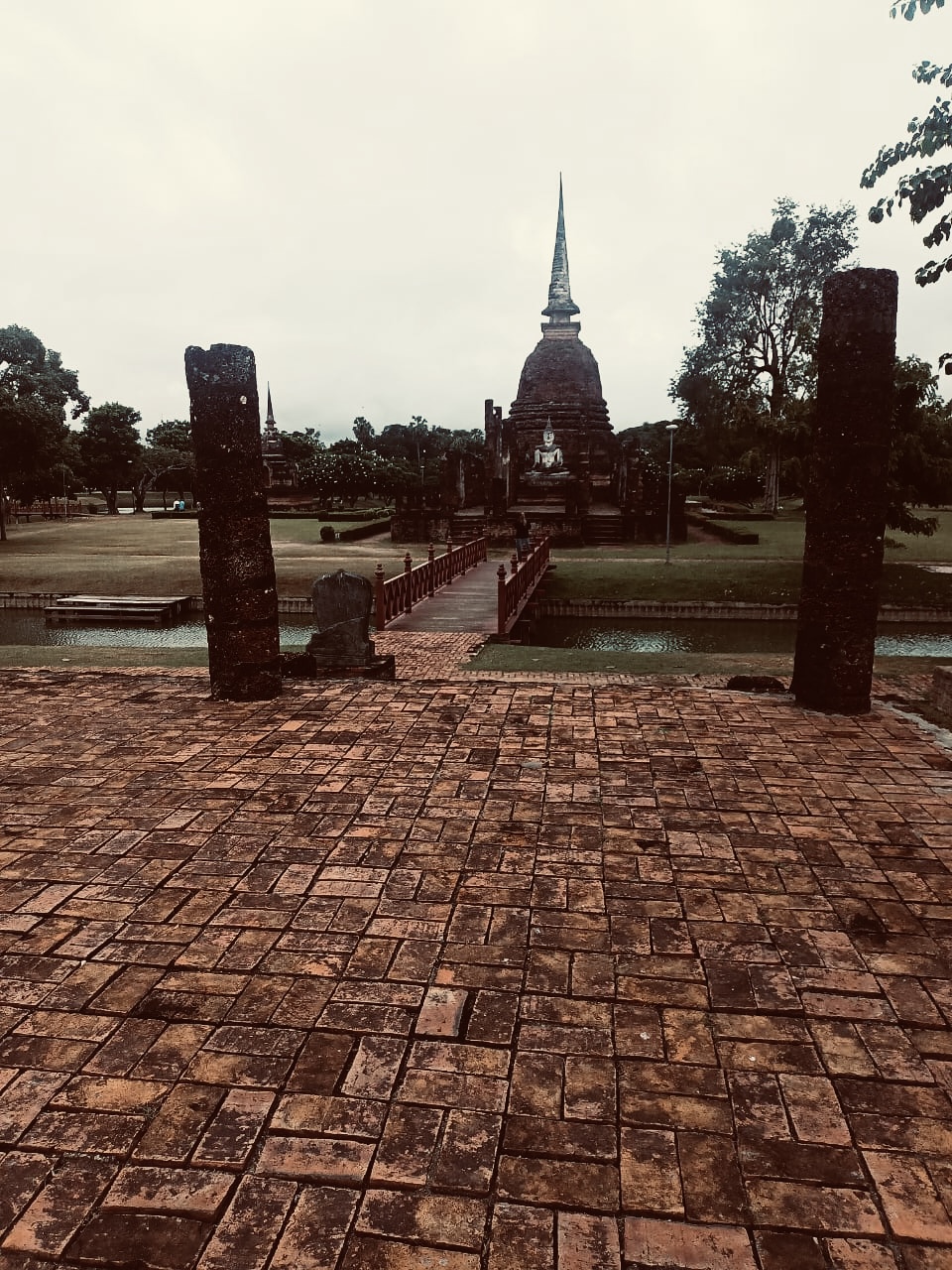 Blick über Ziegelweg und Brücke auf Tempelruinen mit Buddha-Statue im historischen Park von Sukhothai, Thailand