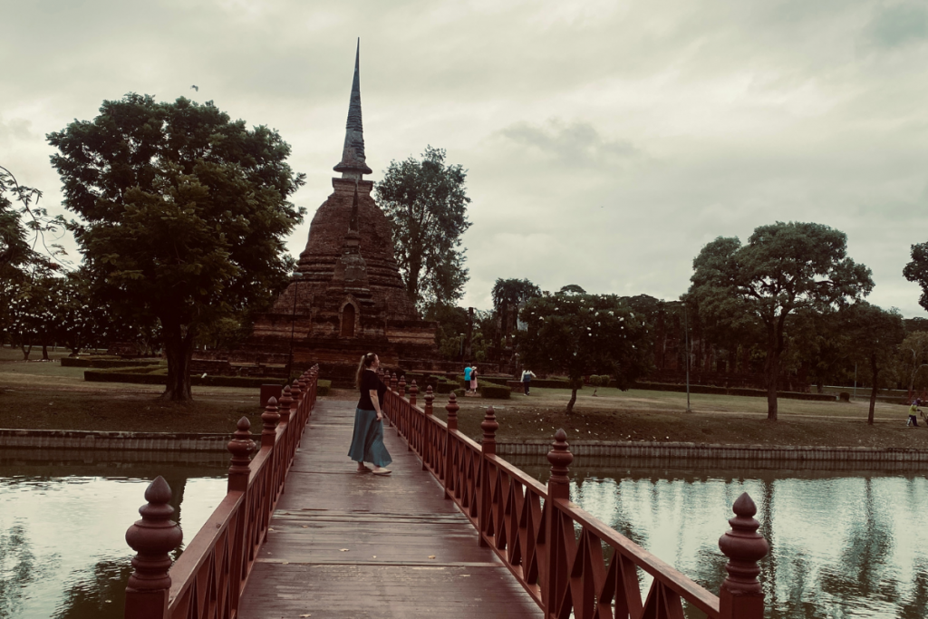 Arbeiten als digitaler Nomade in Sukhothai Thailand - Bensfar Digitale Positionierung Olga Bensfar steht auf roter Brücke vor alter Tempelanlage im historischen Park von Sukhothai, Thailand