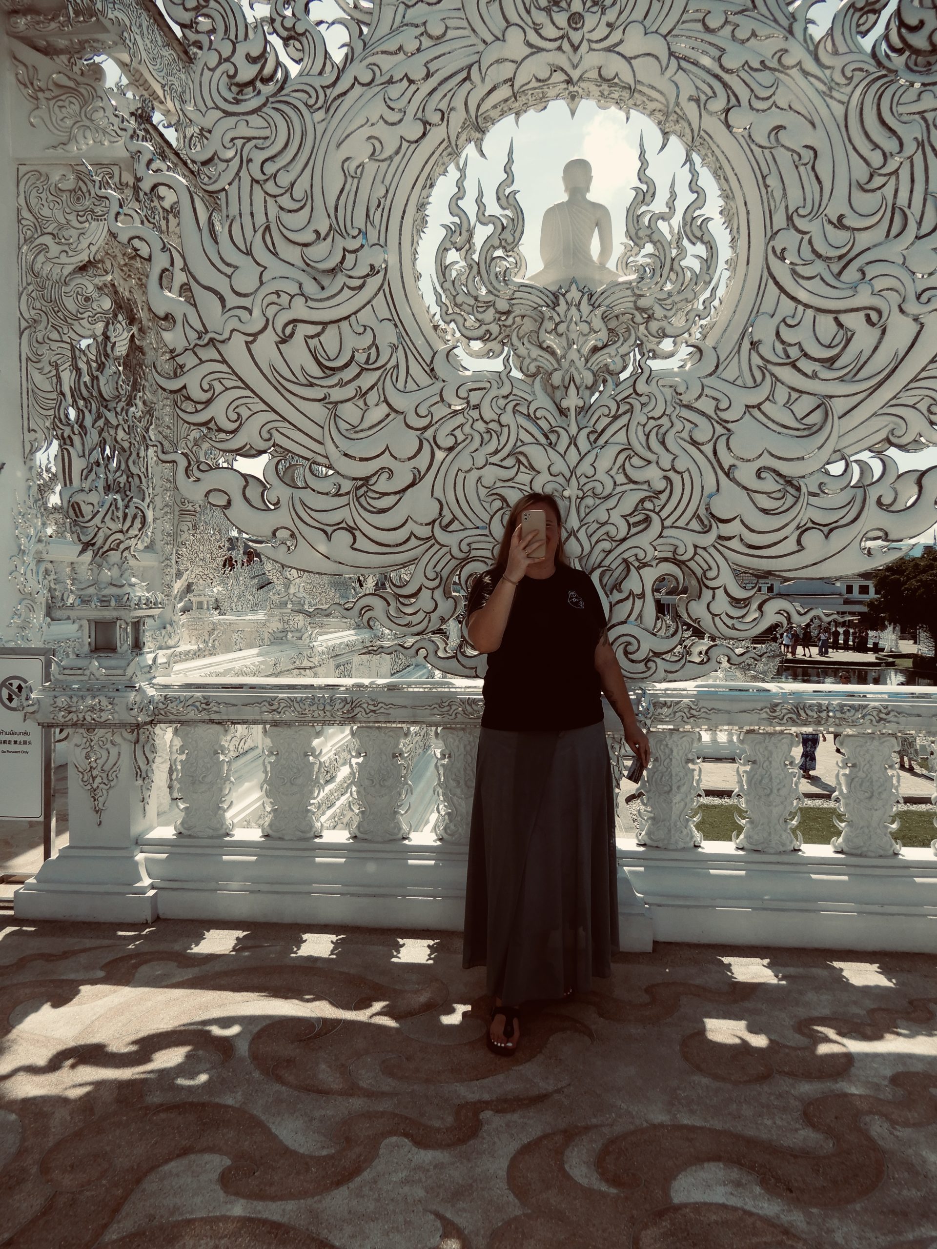 White Temple Chiang Rai – Digitale Freiheit und Inspiration fuer Unternehmerinnen - Bensfar Digitale Positionierung Olga Bensfar steht vor kunstvoll verziertem weißen Tor im White Temple Wat Rong Khun in Chiang Rai, Thailand, mit Buddha-Statue im Hintergrund. ortsunabhaengige Unternehmerin